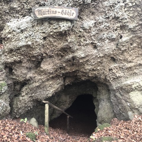 Ice & millstone cave "Martinshöhle" in Hohenfels-Essingen, © Touristik GmbH Gerolsteiner Land, Leonie Post Massive rock face with a small cave entrance inside. A sign with the name ‘Martins-Höhle’ hangs above the cave.