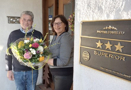 Frank Reuter und Maria Meyer freuen sich über das tolle Klassifizerungsergebnis., © Touristik GmbH Gerolsteiner Land Zwei Personen halten einen Blumestrauß vor der Eingangstür des Landhauses.