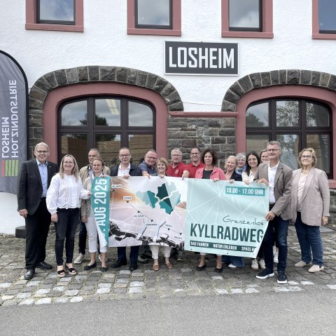 Die Presskonferenz zum Radaktionstag Grenzenlos Kyllradweg 2025 in Losheim., © Gemeinde Hellenthal Vertreter der Ortsgemeinden entlang der Strecke und Politiker stehen dicht beeinander und halten das Veranstaltungsbanner hoch.