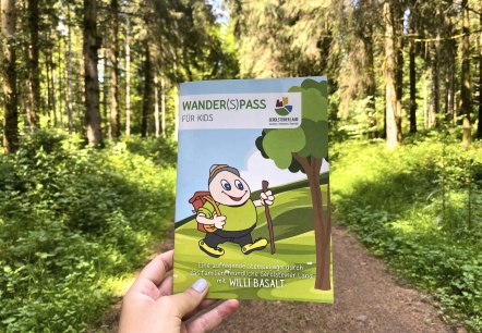 Das Prospekt "Wander(S)pass für Kids" im Wald., © Touristik GmbH Gerolsteiner Land Das Prospekt "Wander(S)pass für Kids" wird hochgehalten mit einem dichten Wald und Waldweg im Hintergrund.