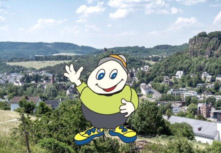 Family-friendly mascot Willi Basalt waves from Gerolstein, © Jochen Hank; Touristik GmbH Gerolsteiner Land Cartoon character Willi Basalt with green shirt, grey trousers and yellow shoes waves. Behind him is a view of the town of Gerolstein with church, buildings and rocks.
