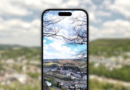 Bilder Instagram, © Touristik GmbH Gerolsteiner Land Vor dem verschwommenen Hintergrund ist ein Handy, welches ein Bild von Gerolstein von oben zeigt.