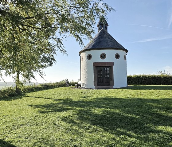 Round chapel on a green meadow, surrounded by trees. The sky is blue and clear., © Touristik GmbH Gerolsteiner Land Round chapel on a green meadow, surrounded by trees. The sky is blue and clear., © Touristik GmbH Gerolsteiner Land