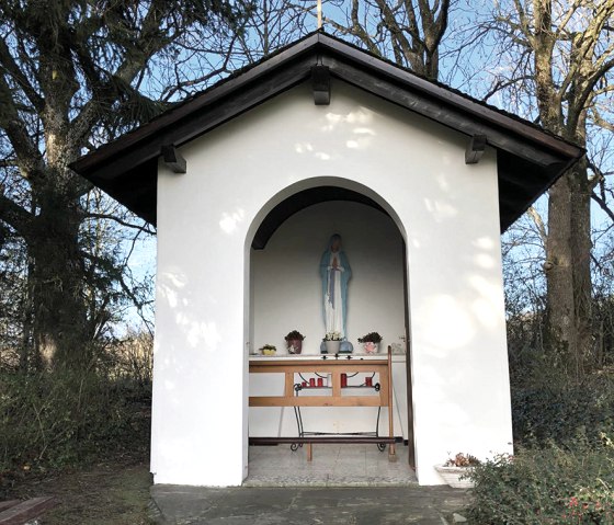 White chapel with a statue of the Virgin Mary and a cross, surrounded by trees. The sky is blue and clear., © Touristik GmbH Gerolsteiner Land White chapel with a statue of the Virgin Mary and a cross, surrounded by trees. The sky is blue and clear., © Touristik GmbH Gerolsteiner Land