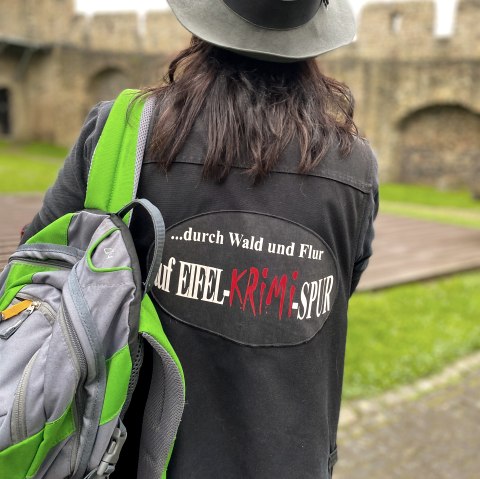 Crime scene lady in front of the Hillesheim town wall, © Touristik GmbH Gerolsteiner Land, Gianne Kern View of the back of a woman wearing a hat and a printed jacket and carrying a green rucksack over one shoulder. In front of her is the Hillesheim town wall.