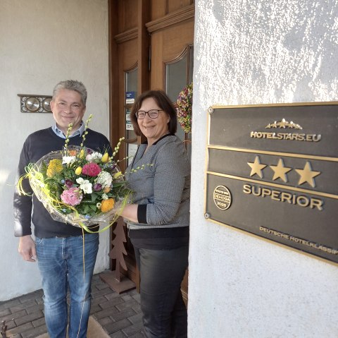 Frank Reuter und Maria Meyer freuen sich über das tolle Klassifizerungsergebnis., © Touristik GmbH Gerolsteiner Land Zwei Personen halten einen Blumestrauß vor der Eingangstür des Landhauses.