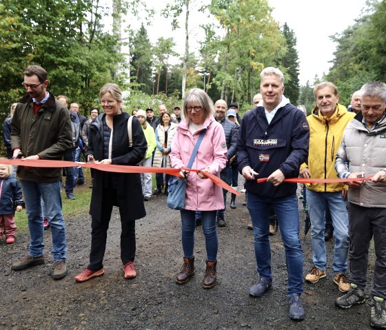 Landtagsabgeordneter Jens Jenssen, Landrätin Julia Gieseking, Verbandsgemeindebürgermeister Hans Peter Böffgen, Geschäftsführer Frank Reuter und Stadtbürgermeisterin Steffi Lorisch durchschneiden gemeinsam das rote Band und geben damit die Waldkugelbahn offiziell frei., © Thomas Langens Fünf Personen mit Scheren in den Händen durchschneiden gleichzeitig ein rotes Band. Hinter ihnen zahlreiche Zuschauer und Kinder.