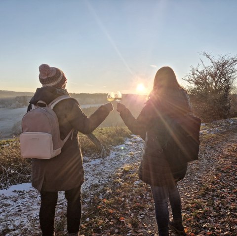 Wein mit Aussicht, Wanderer mit Weingläsern bei Sonnenuntergang
, © Touristik GmbH Gerolsteiner Land, Esther Erharter Zwei Personen stehen auf einem mit etwas Schnee bedeckten Wanderweg und stoßen mit zwei Weingläsern an. Im Hintergrund geht die Sonne hinter Feldern unter.