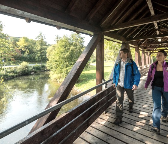 Wanderer überqueren die Kyll im Kurpark Gerolstein über die Holzbrücke.
, © Eifel Tourismus GmbH, Dominik Ketz Mann und Frau in Wanderkleidung gehen über eine Holzbrücke über den Fluss Kyll im Kurpark Gerolstein.