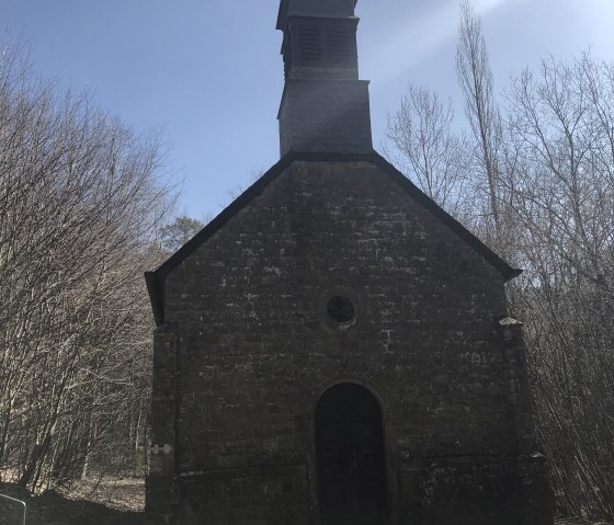 Die Büschkapelle in Gerolstein, aus Stein gebaut, steht vor einem klaren blauen Himmel. Bäume umgeben die Kapelle, die einen kleinen Turm hat., © Touristik GmbH Gerolsteiner Land, Leonie Post Die Büschkapelle in Gerolstein, aus Stein gebaut, steht vor einem klaren blauen Himmel. Bäume umgeben die Kapelle, die einen kleinen Turm hat., © Touristik GmbH Gerolsteiner Land, Leonie Post