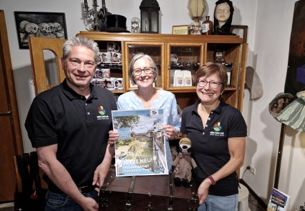 De schatkist wacht in de boekhandel van Lesezeichen aan het einde van het detectivetraject op nieuwsgierige speurders., © Touristik GmbH Gerolsteiner Land Drie lachende mensen staan in een kleine kamer voor een houten kast en houden een grote schatkist en een poster over het detectivetraject omhoog.