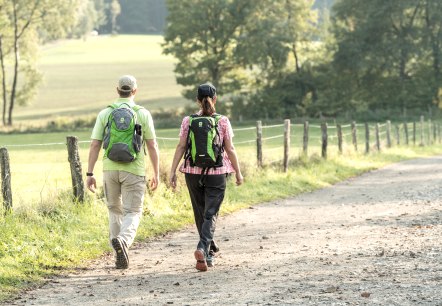 Two people are walking along a rural path in the Eifel. They are carrying rucksacks and are surrounded by green countryside., © Eifel Tourismus GmbH, Dominik Ketz Two people are walking along a rural path in the Eifel. They are carrying rucksacks and are surrounded by green countryside., © Eifel Tourismus GmbH, Dominik Ketz