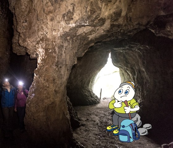 Family-friendly mascot Willi Basalt visits the Buchenloch Cave in Gerolstein, © Eifel Touristik GmbH, Dominik Ketz; Touristik GmbH Gerolsteiner Land Cartoon character Willi Basalt with green shirt, grey trousers and yellow shoes sits on a stone with rucksack and walking stick. Surrounding him are the rock faces of the Buchenloch Cave and two other visitors.