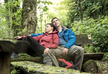 Wandelaars pauzeren in het bos.
, © Eifel Tourismus GmbH, Dominik Ketz Een man en een vrouw in wandelkleding zitten ontspannen tegen elkaar aan op een bankje midden in het bos.