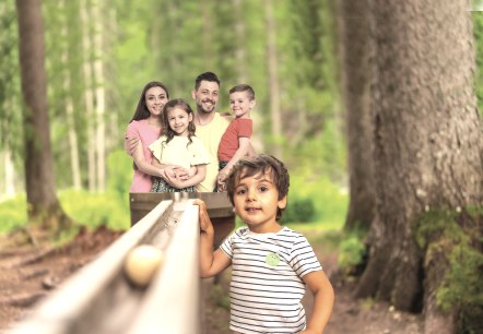Familie an der Waldkugelbahn, © Kurt Daun Eltern mit zwei Kindern auf dem Arm stehen hinter einer Hoz-Kugelbahn und ein Kind steht daneben im Wald.