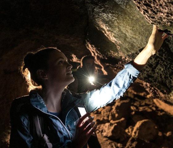 Een persoon in een grot verlicht het plafond met een fakkel, terwijl een andere persoon op de achtergrond ook een lamp vasthoudt., © Eifel Tourismus GmbH, Dominik Ketz Een persoon in een grot verlicht het plafond met een fakkel, terwijl een andere persoon op de achtergrond ook een lamp vasthoudt., © Eifel Tourismus GmbH, Dominik Ketz