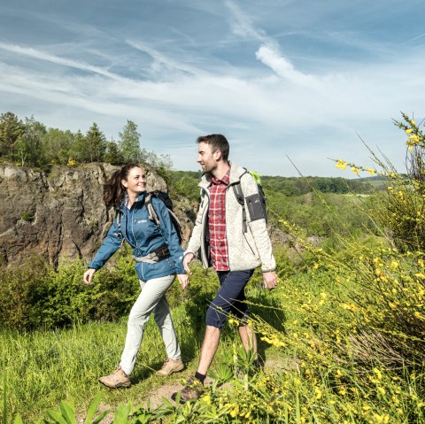 Wanderer im Vulkangarten Steffeln vor dem Vulkan Steffelnkopf., © Eifel Tourismus GmbH, Dominik Ketz Zwei Wanderer inmitten der Natur auf einem Wanderweg. Im Hintergrund ist eine große Felswand zu sehen. Hierbei handelt es sich um den Vulkan Steffelnkopf.
