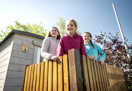 Drie meisjes bezoeken de schaapskooien in het Kasselburger Adelaar- en Wolfspark.
, © Eifel Tourismus GmbH, Dominik Ketz Drie jonge meisjes staan bij een houten hek en kijken er blij overheen. Achter hen staat een klein houten huisje.