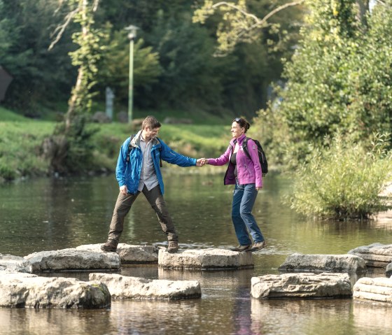 A couple crosses stepping stones in the Kyll near Gerolstein. They are holding hands and wearing outdoor clothing., © Eifel Tourismus GmbH, Dominik Ketz A couple crosses stepping stones in the Kyll near Gerolstein. They are holding hands and wearing outdoor clothing., © Eifel Tourismus GmbH, Dominik Ketz