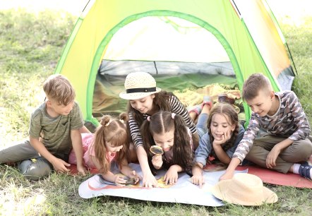 Sechs Kinder untersuchen eine Landkarte, die vor ihnen liegt., © Adobe Stocks Sechs Kinder liegen und sitzen dicht beieinander vor einem bunten Zelt auf einer Wiese und schauen auf eine Landkarte, die vor ihnen liegt. Manche Kinder zeigen auf die Karte und ein Kind schaut genauer mit einer Lupe.