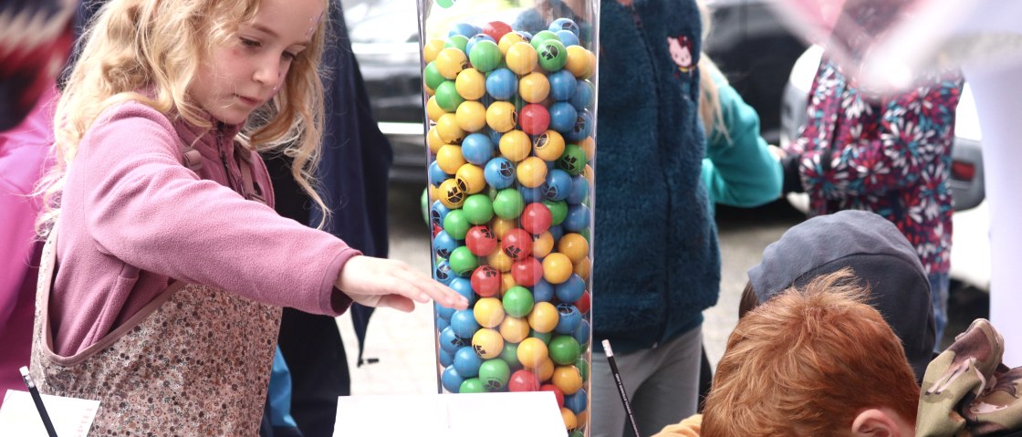 Das Gewinnspiel der Touristik GmbH Gerolsteiner Land lud zahlreiche Besucher zum Schätzen ein.
, © Thomas Langens Mehrere Kinder stehen um ein Glasgefäß herum, welches mit zahlreichen bunten Kugeln gefüllt ist, und schreiben auf Blättern.