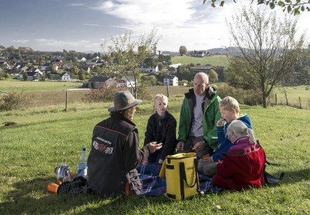 Familie en vrouwelijke detective op een picknick
, © Kappest Twee volwassenen en twee kinderen zitten op een deken in een ruime weide naast een boom. Ze hebben proviand in rugzakken.