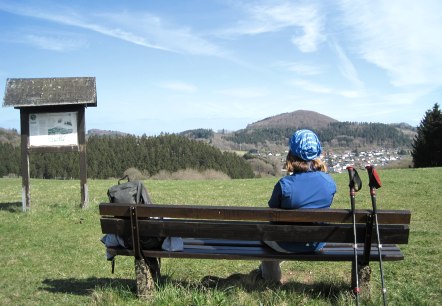 Een persoon zit op een bankje en kijkt naar de Nerother Kopf. Wandelstokken leunen tegen het bankje, een rugzak ligt ernaast. De lucht is blauw., © Touristik GmbH Gerolsteiner Land, Ute Klinkhammer Een persoon zit op een bankje en kijkt naar de Nerother Kopf. Wandelstokken leunen tegen het bankje, een rugzak ligt ernaast. De lucht is blauw., © Touristik GmbH Gerolsteiner Land, Ute Klinkhammer