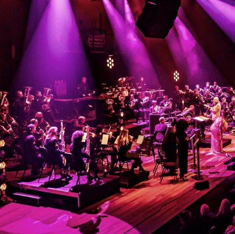Das Bild zeigt ein Orchester bei einer Aufführung auf einer Bühne, die in einem lebhaften Magenta-Ton beleuchtet ist. Das Orchester besteht aus zahlreichen Musikern, die verschiedene Instrumente spielen, darunter Blasinstrumente, Streichinstrumente und Schlagzeug. Ein Dirigent steht vor dem Orchester, und eine Sängerin in einem glitzernden Kleid steht ebenfalls auf der Bühne. Im Vordergrund ist ein Teil des Publikums zu sehen, das der Aufführung zusieht. Die Beleuchtung trägt zur dramatischen Atmosphäre bei., © Touristik GmbH Gerolsteiner Land Das Bild zeigt ein Orchester bei einer Aufführung auf einer Bühne, die in einem lebhaften Magenta-Ton beleuchtet ist. Das Orchester besteht aus zahlreichen Musikern, die verschiedene Instrumente spielen, darunter Blasinstrumente, Streichinstrumente und Schlagzeug. Ein Dirigent steht vor dem Orchester, und eine Sängerin in einem glitzernden Kleid steht ebenfalls auf der Bühne. Im Vordergrund ist ein Teil des Publikums zu sehen, das der Aufführung zusieht. Die Beleuchtung trägt zur dramatischen Atmosphäre bei., © Touristik GmbH Gerolsteiner Land