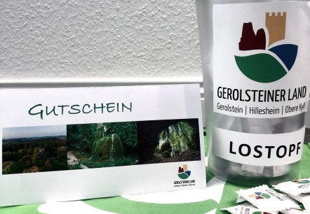 , © Touristik GmbH Gerolsteiner Land A lottery pot with the logo of the holiday region stands on a table, next to it the travel voucher to be won and in front of it some dextrose.