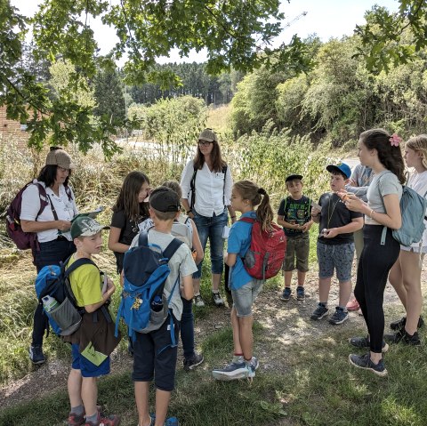 Kindergruppe beim Kinder-Krimiprogramm mit zwei Krimi-Damen, © Touristik GmbH Gerolsteiner Land Eine Gruppe von Kindern steht zusammen auf einem Pfad vor zwei erwachsenen Frauen, die beide Detektivhüte tragen und den Kindern etwas erzählen.