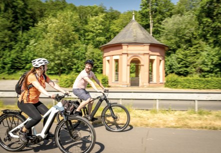 Twee fietsers op het Kyll-fietspad bij de Lindenquelle bij Birresborn.
, © Eifel Tourismus GmbH, Dominik Ketz Een man en een vrouw fietsen op e-bikes langs een fietspad langs de grote bron van de Lindenquelle bij Birresborn.