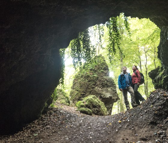 Two people are standing in a cave and looking out. The cave is surrounded by green plants and trees can be seen in the background., © Eifel Tourismus GmbH, Dominik Ketz Two people are standing in a cave and looking out. The cave is surrounded by green plants and trees can be seen in the background., © Eifel Tourismus GmbH, Dominik Ketz