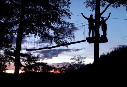 Zwei Personen bei Night-Event im EifelAdventures Berlingen auf der Ziplining-Plattform.
, © EifelAdventures Berlingen Zwei Personen stehen in der Dunkelheit auf einer Plattform an einem Baum und sind an Seilen gesichert. Sie Stehen mitten in einem Kletterparcours und hinter ihnen geht die Sonne unter.