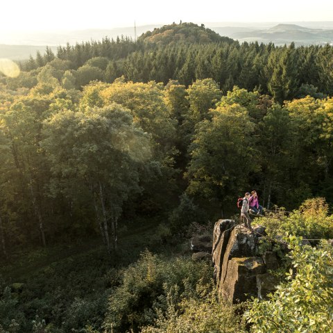 Zwei Personen stehen auf einem Felsen, umgeben von einem dichten Wald. Im Hintergrund sind Hügel und ein weiter Blick über die Landschaft zu sehen., © Eifel Tourismus GmbH, Dominik Ketz Zwei Personen stehen auf einem Felsen mit Blick auf einen Wald und Hügel im Hintergrund.