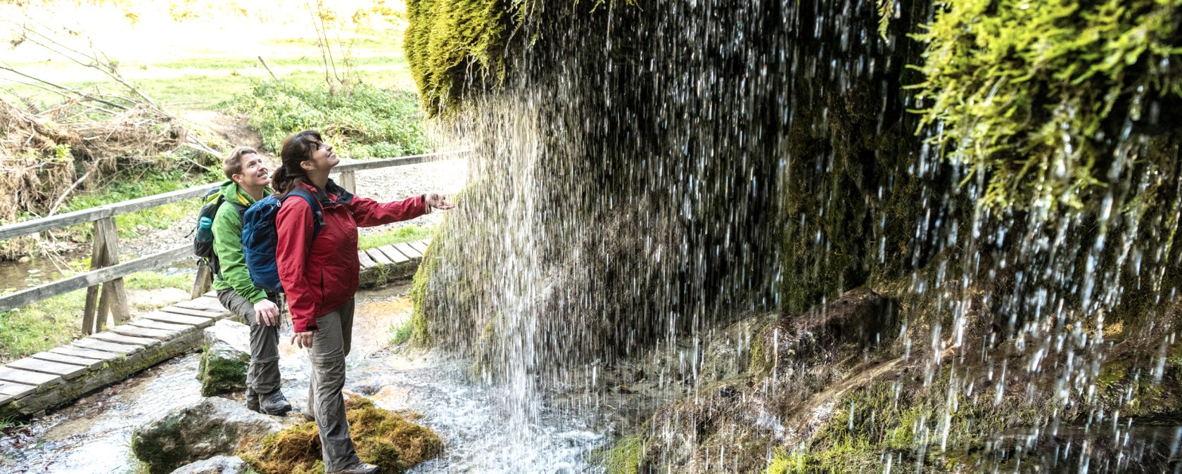 Two people look at the moss-covered waterfall in the green forest landscape. A wooden footbridge leads over the stream., © Eifel Tourismus GmbH - Dominik Ketz Two people look at the moss-covered waterfall in the green forest landscape. A wooden footbridge leads over the stream., © Eifel Tourismus GmbH - Dominik Ketz