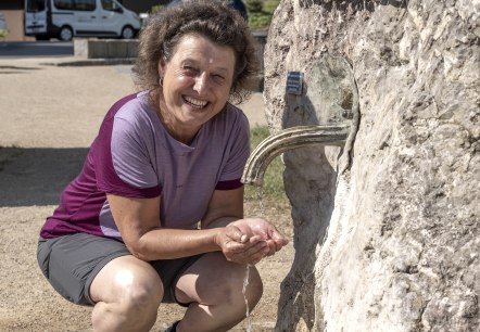 Eine Wanderin erfreut sich am frischen Mineralwasser der Helenenquelle., © Jochen Hank Eine Frau kmiet menem einem Felsen aus dem ein Hahn, aus welchem Wasser herausströmt. Die Frau fängt das Wasser mit den Händen auf.