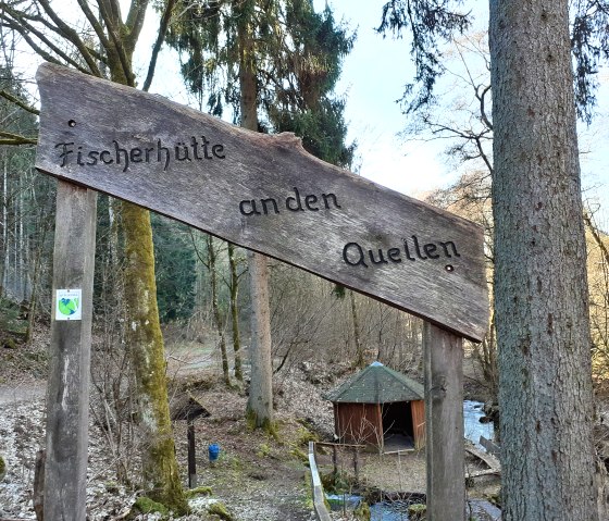 Holzschild mit der Aufschrift 'Fischerhütte an den Quellen' im Wald, im Hintergrund eine kleine Hütte und ein Bach., © Touristik GmbH Gerolsteiner Land, Ute Klinkhammer Holzschild mit der Aufschrift 'Fischerhütte an den Quellen' im Wald, im Hintergrund eine kleine Hütte und ein Bach., © Touristik GmbH Gerolsteiner Land, Ute Klinkhammer