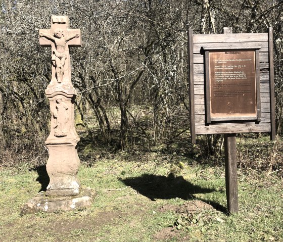 Ein Steinkreuz mit Christusfigur steht neben einer hölzernen Infotafel im Wald. Der Boden ist grasbewachsen, die Bäume sind kahl., © Touristik GmbH Gerolsteiner Land, Leonie Post Ein Steinkreuz mit Christusfigur steht neben einer hölzernen Infotafel im Wald. Der Boden ist grasbewachsen, die Bäume sind kahl., © Touristik GmbH Gerolsteiner Land, Leonie Post
