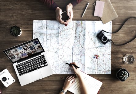 Urlaubsplanung, © pixabay Großer Holztsich, an dem zwei Personen sitzen. Darauf eine Landkarte, Kameras, Zettel, Stifte und ein Laptop.