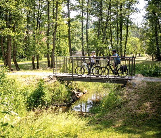 Zwei Radfahrer im Kurpark Stadtkyll
, © Eifel Tourismus GmbH, Dominik Ketz Zwei Personen fahren auf Fahrrädern über eine Brücke in einer grün blühenden Parkanlage.