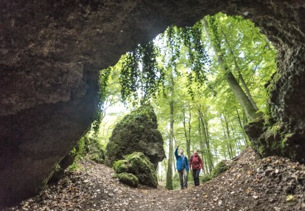Hikers in front of the Birresborn ice caves., © Eifel Tourismus GmbH, Dominik Ketz Two people walk towards a cave entrance made of stone and point to the rocks around them. The forest in front of the caves is full of tall green blossoming trees.