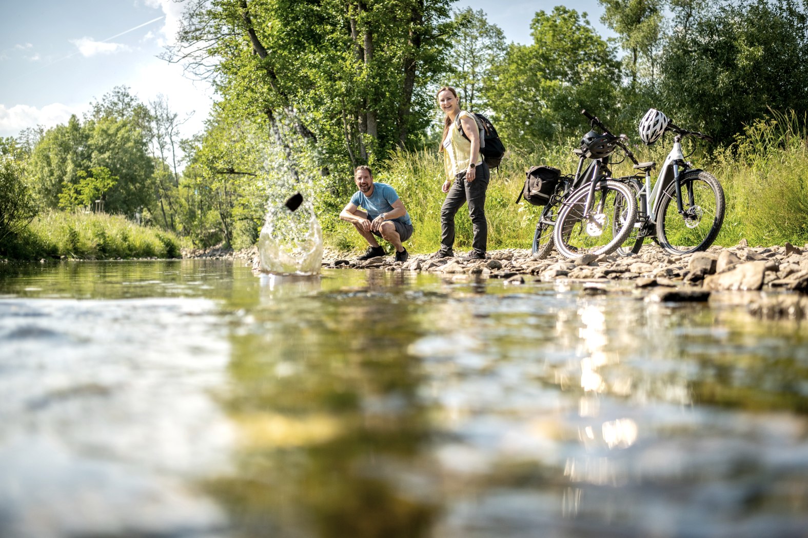 Radtour Rast im Kurpark Stadtkyll an der Kyll.
, © Eifel Tourismus GmbH, Dominik Ketz Zwei Personen stehen neben Ihren Fahrrädern am Ufer eines Flusses und werfen Steine über die Wasseroberfläche.