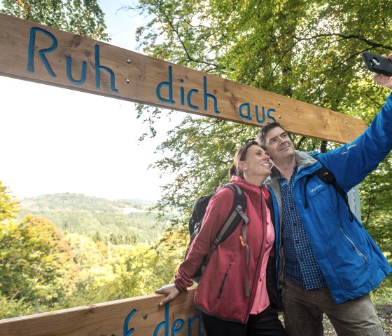 Ein Paar macht ein Selfie unter einem Holzschild mit der Aufschrift 'Ruh dich aus' in einem grünen Waldgebiet., © Eifel Tourismus GmbH, Dominik Ketz Ein Paar macht ein Selfie unter einem Holzschild mit der Aufschrift 'Ruh dich aus' in einem grünen Waldgebiet., © Eifel Tourismus GmbH, Dominik Ketz