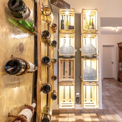 Weinregal in Ferienwohnung des Hof Grindelborn
, © Hof Grindelborn Holzplatten an einer Wand mit Weinflaschen, die durch Seile daran befestigt sind. Daneben Weinregale aus Holzkisten mit weiteren Flaschen und Schieferplatten mit darauf geschriebenen Sortiment.