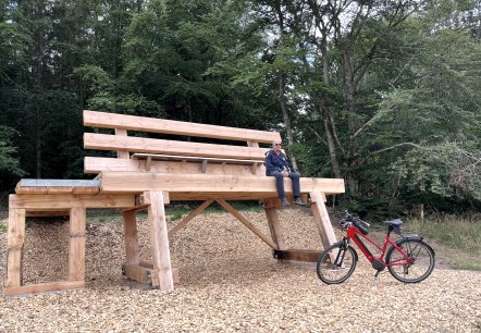 A huge wooden bench in the forest with a person on it. A red bicycle stands next to it on wood chips., © Touristik GmbH Gerolsteiner Land A huge wooden bench in the forest with a person on it. A red bicycle stands next to it on wood chips., © Touristik GmbH Gerolsteiner Land