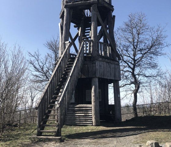 Ein hölzerner Aussichtsturm mit Treppe und Geländer steht auf einem Hügel, umgeben von kahlen Bäumen und blauem Himmel., © Touristik GmbH Gerolsteiner Land, Leonie Post Ein hölzerner Aussichtsturm mit Treppe und Geländer steht auf einem Hügel, umgeben von kahlen Bäumen und blauem Himmel., © Touristik GmbH Gerolsteiner Land, Leonie Post