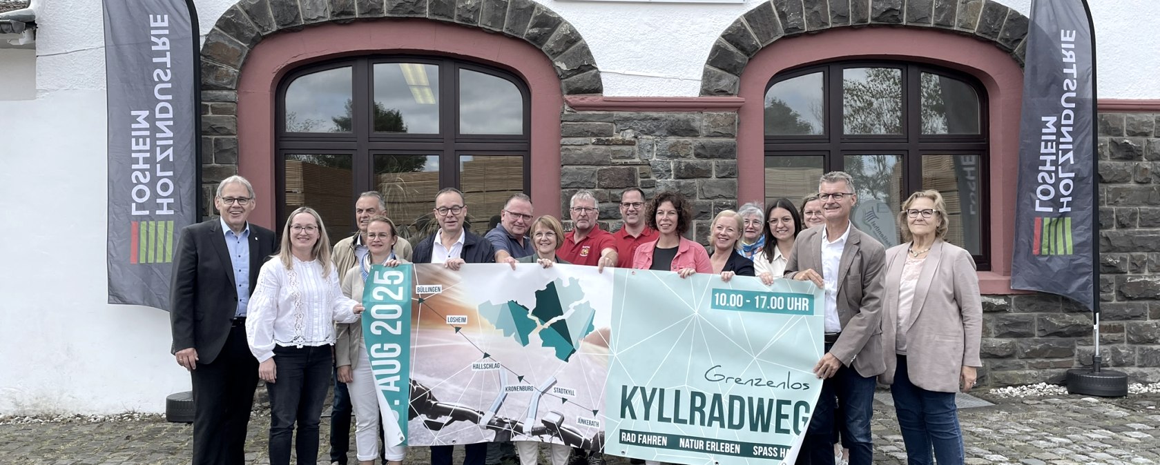 Die Presskonferenz zum Radaktionstag Grenzenlos Kyllradweg 2025 in Losheim., © Gemeinde Hellenthal Vertreter der Ortsgemeinden entlang der Strecke und Politiker stehen dicht beeinander und halten das Veranstaltungsbanner hoch.