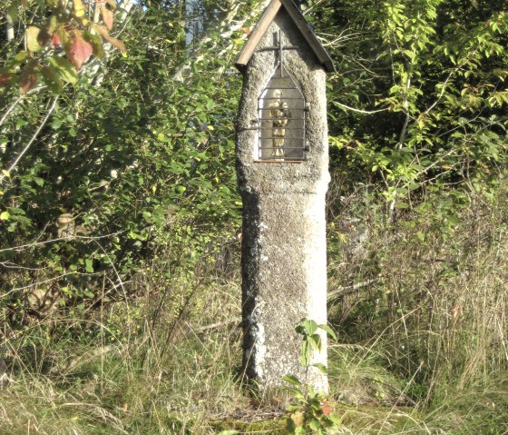 A stone wayside cross with a small roof and lattice stands in the middle of grass and trees., © Touristik GmbH Gerolsteiner Land A stone wayside cross with a small roof and lattice stands in the middle of grass and trees., © Touristik GmbH Gerolsteiner Land