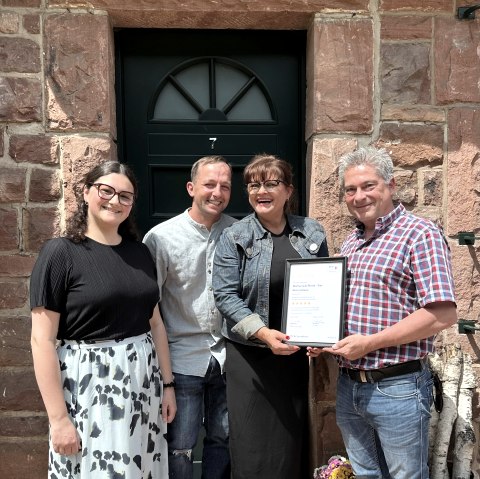 Erfolgreiche Klassifizierung der Ferienwohnungen im Hof Grindelborn, Mürlenbach., © Hof Grindelborn Vier erfreute und lachende Personen stehen vor einer Eingangstür und halten Stolz die Urkunde für die Klassifizierung in den Händen.