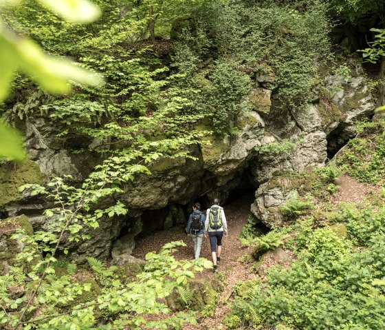 Zwei Wanderer betreten eine von Pflanzen umgebene Höhle im Wald auf dem Vulkanpfad in Gerolstein-Roth., © Eifel Tourismus GmbH, Dominik Ketz Zwei Wanderer betreten eine von Pflanzen umgebene Höhle im Wald auf dem Vulkanpfad in Gerolstein-Roth., © Eifel Tourismus GmbH, Dominik Ketz